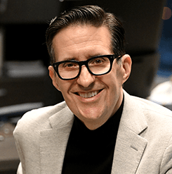 smiling man with dark hair and glasses and a light coloured suit